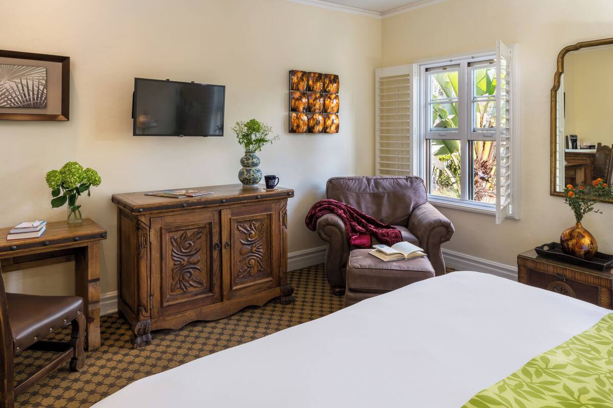 A bedroom with a bed, a wooden dresser, a leather armchair, a television, and a window with blinds.
