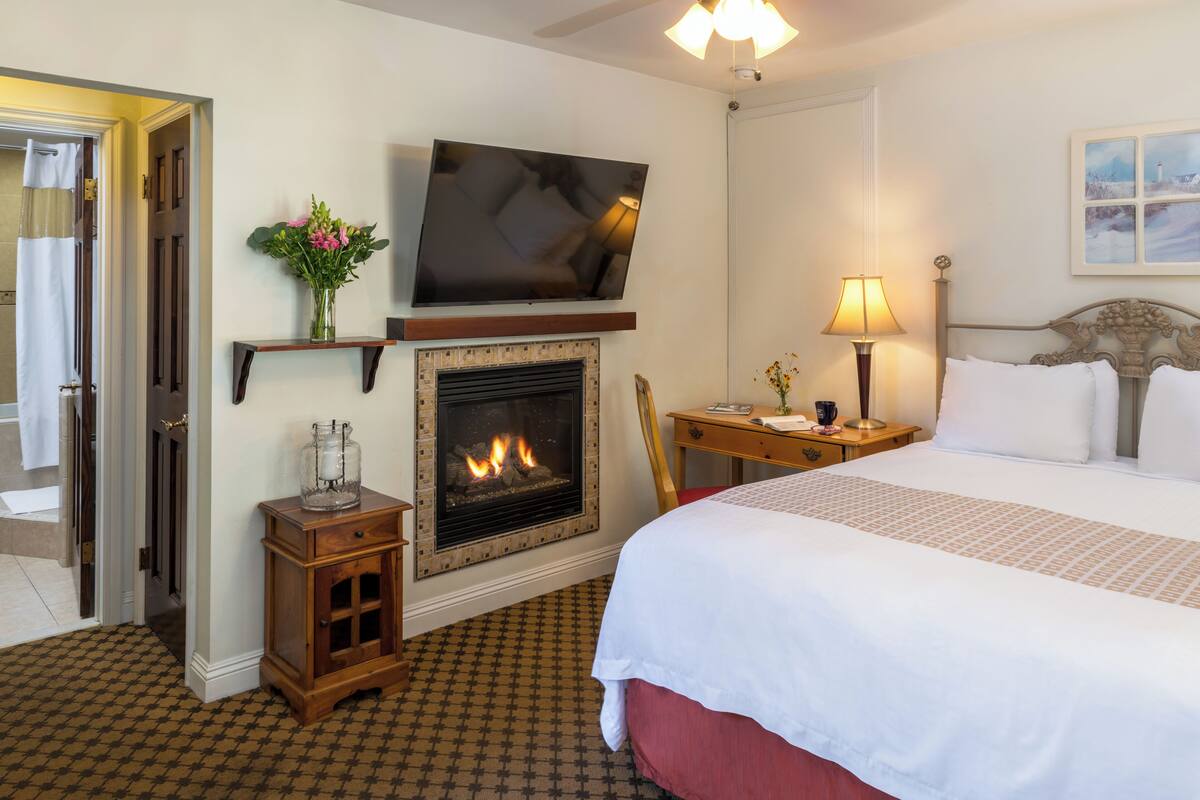 A hotel room with a fireplace, a bed, a TV, a bedside table with a lamp, and a painting on the wall.