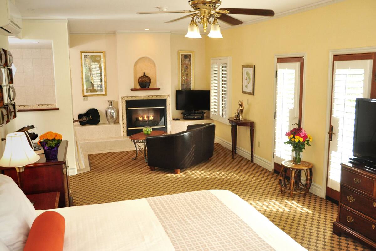 A hotel room with a bed, a television, a fireplace, a ceiling fan, and a window with blinds.