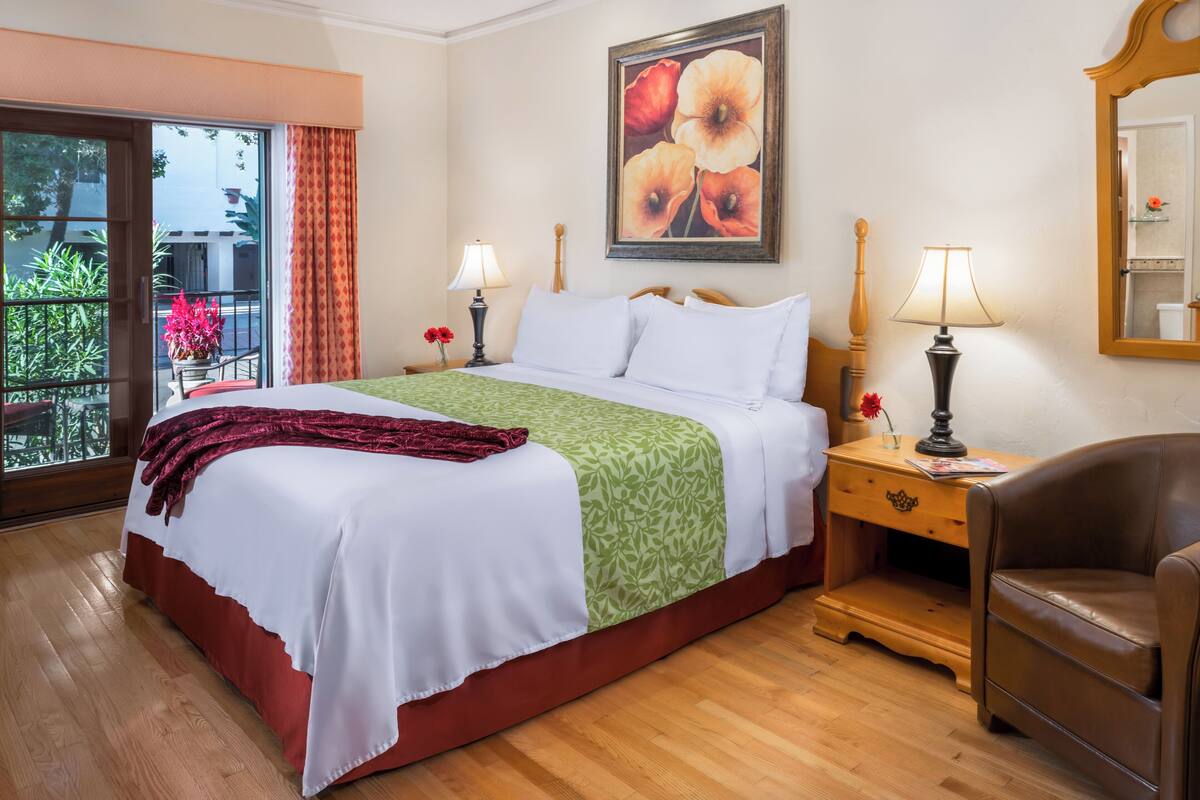 A hotel room with a large bed, two bedside tables with lamps, a wooden chair, a balcony with red curtains, and a painting of flowers on the wall.