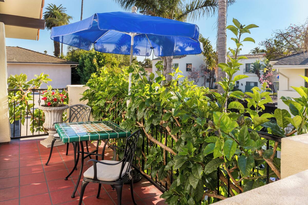 A patio with a table and chairs, a blue umbrella, and lush greenery.