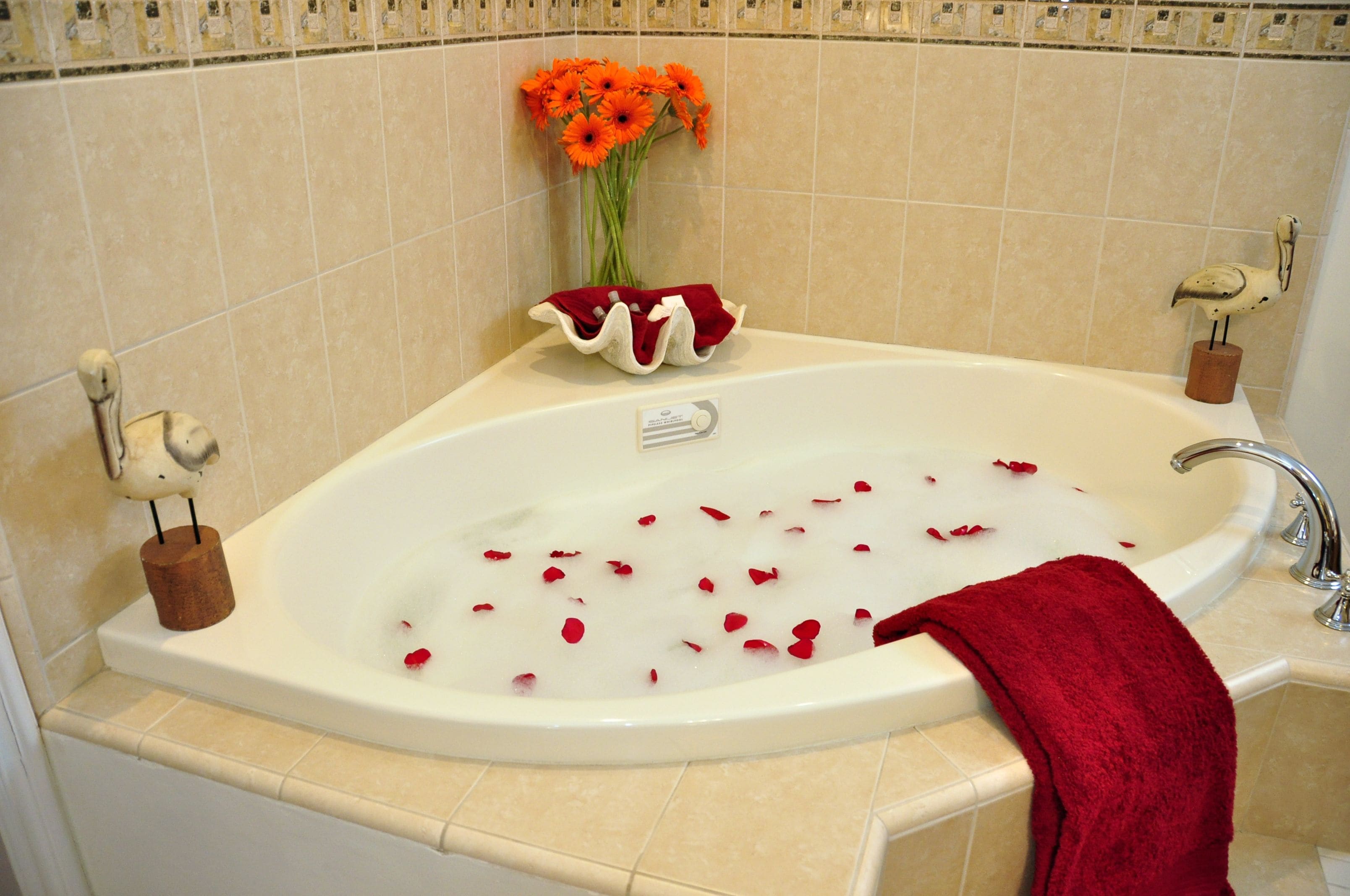 A bathtub with rose petals, a red towel, and a vase with orange flowers.