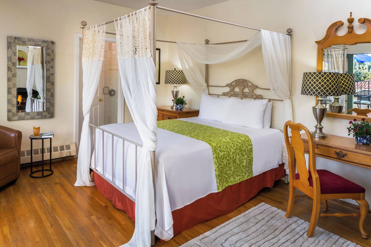 A four-poster canopy bed with a green throw blanket, a wooden desk with a chair, and a mirror on the wall.