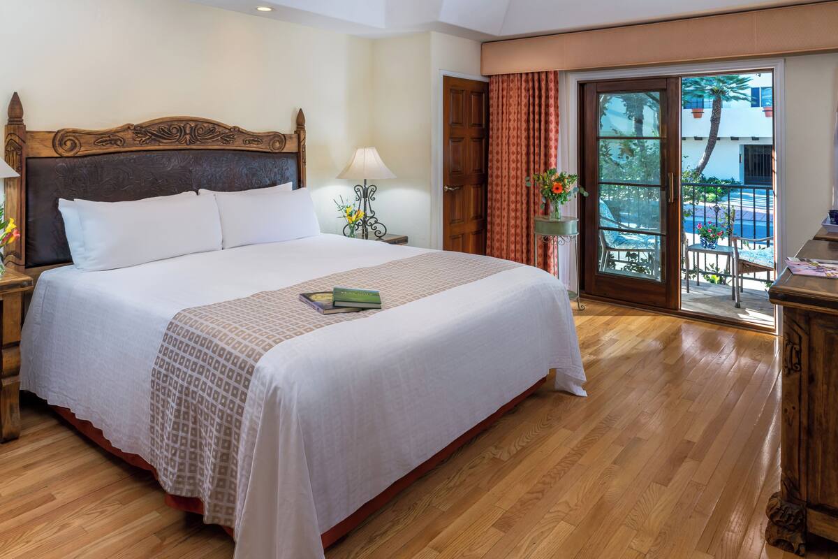 A hotel room with a large bed, wooden headboard, two bedside tables with lamps, a wooden nightstand, and a view of the outdoors through a glass door.