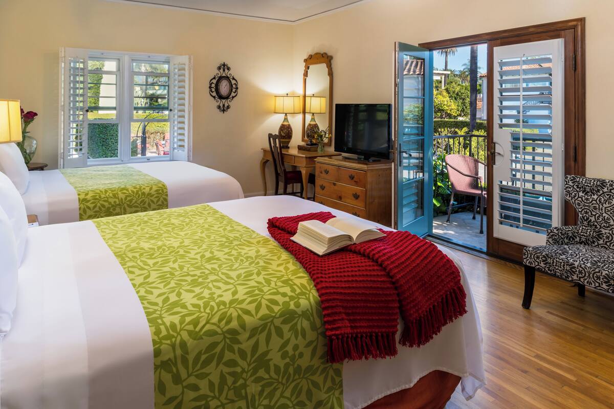A hotel room with two beds, a TV, a desk with a chair, and a balcony with a chair.