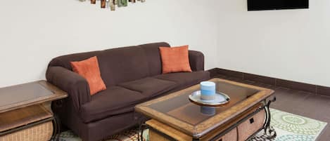 A living room with a brown sofa, orange cushions, a glass-top coffee table, wicker baskets, and a patterned rug.