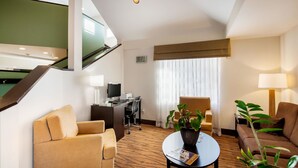 A hotel room with a sofa, armchairs, a desk with a computer, a TV, and a plant.