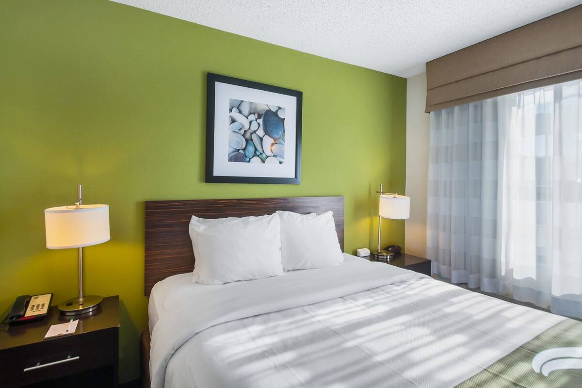 A hotel room with a bed, two bedside lamps, a nightstand with a phone, a framed picture on the wall, and a window with sheer curtains.