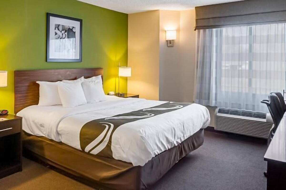 A hotel room with a bed, bedside lamps, a desk, a chair, a window with curtains, and a wall-mounted picture.