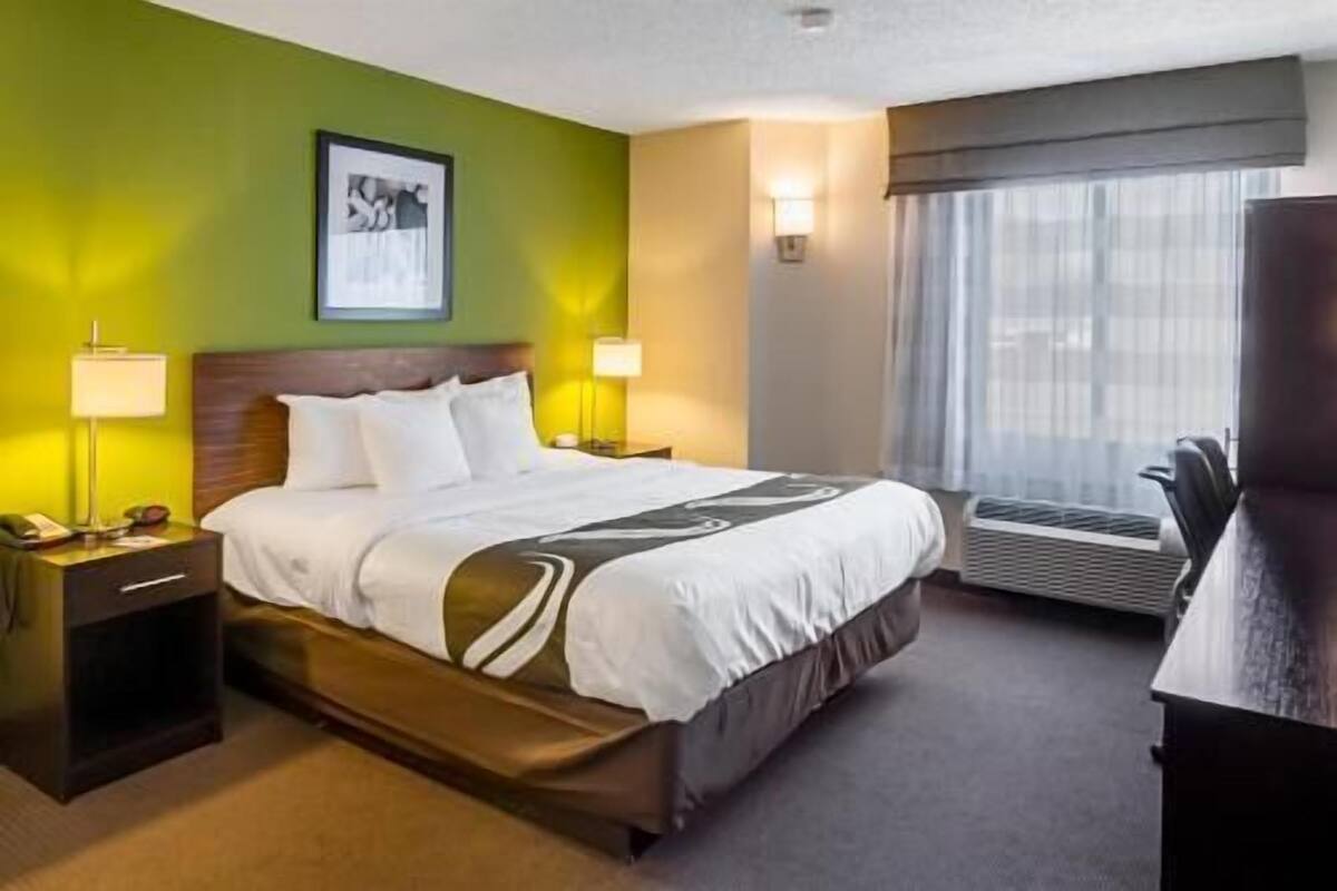 A hotel room with a bed, bedside lamps, a framed picture on the wall, a window with curtains, and an air conditioning unit.