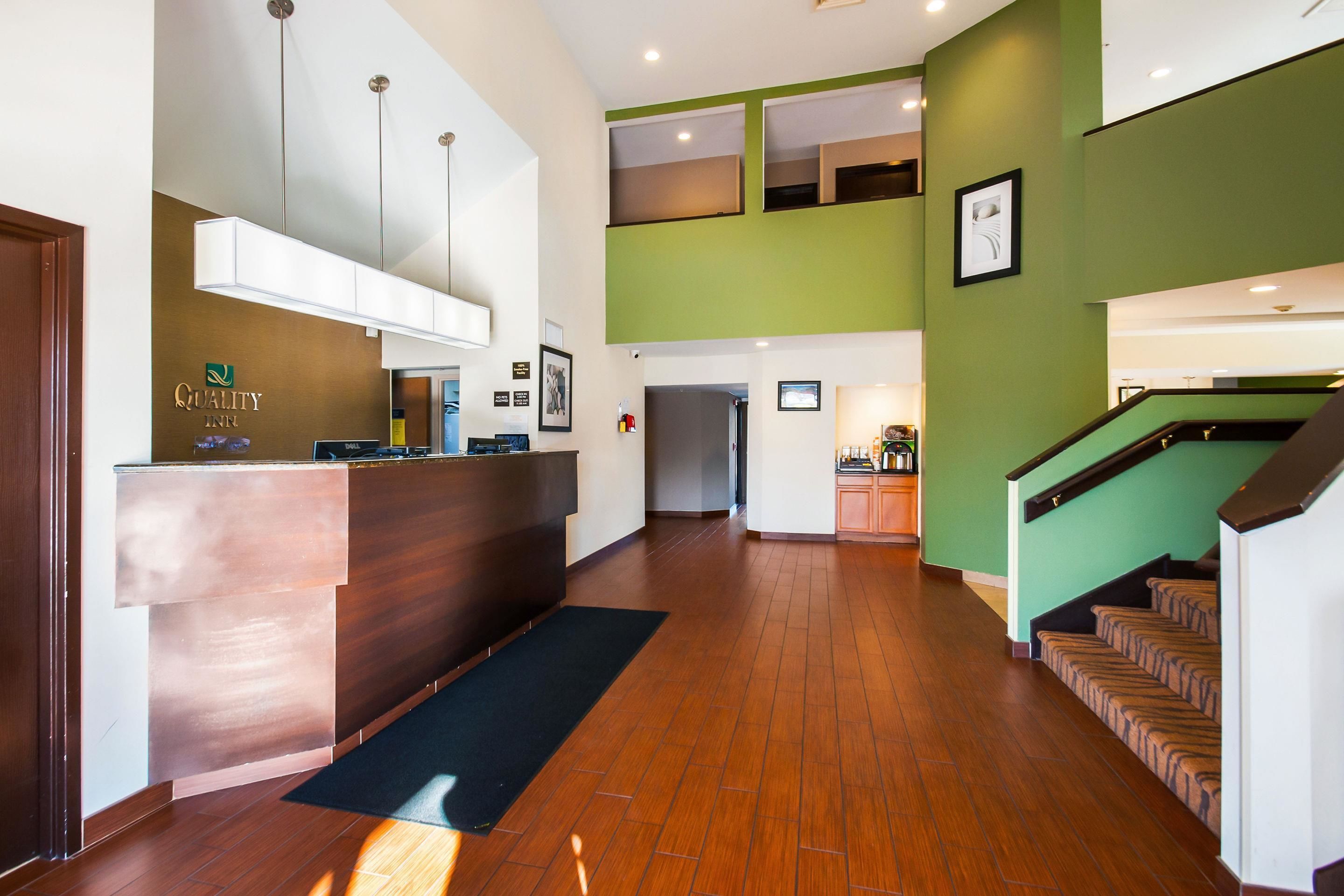 A hotel reception area with a wooden reception desk, a black mat, a staircase, and a green wall.