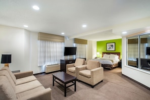 A hotel room with a sofa, armchairs, a TV, a bed, and a large window.