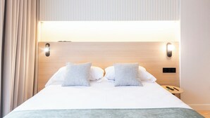 Una cama bien hecha con dos almohadas, una mesita de noche con lámpara y un enchufe en la pared.