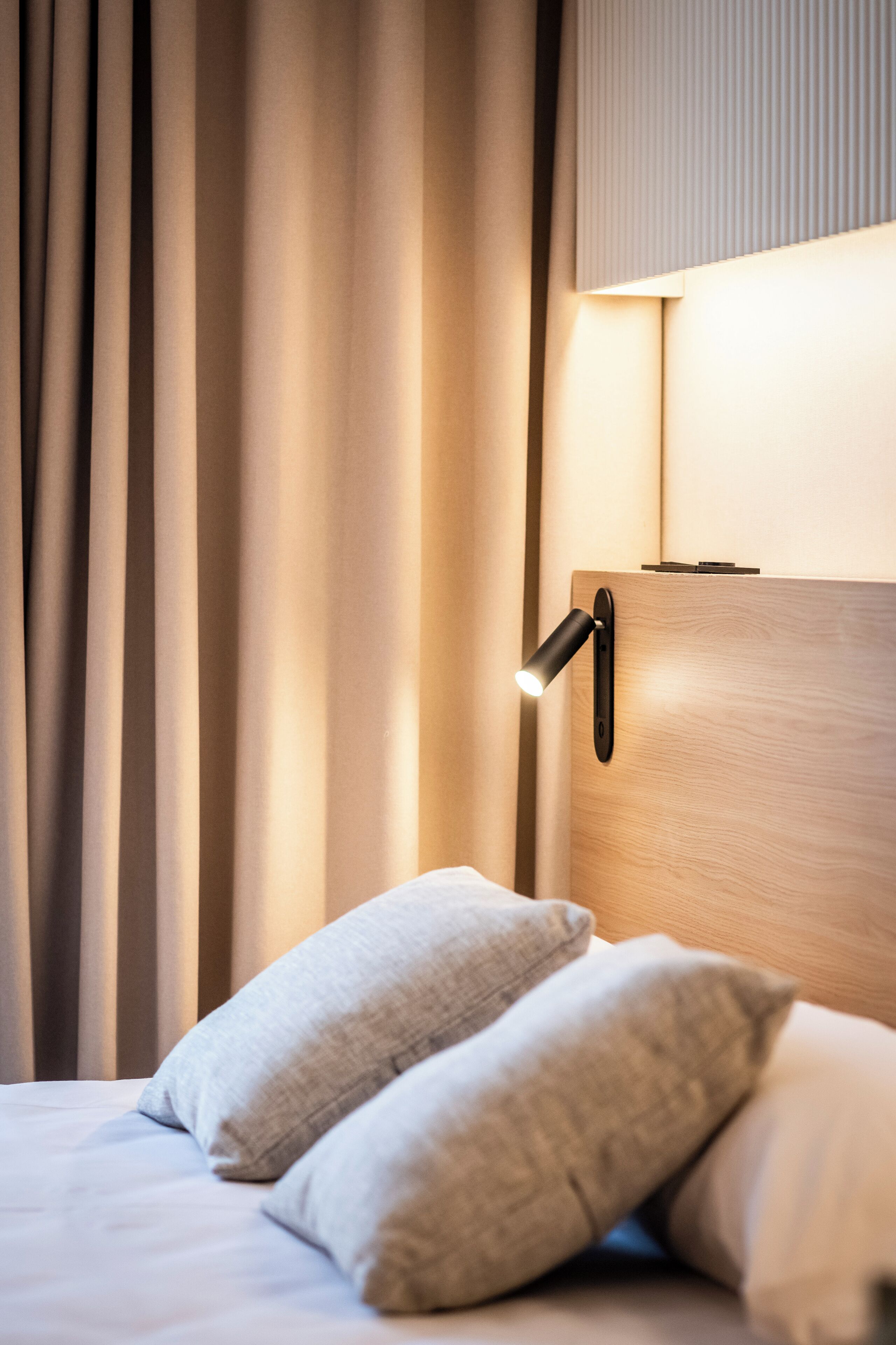 Hotellihuone, jossa on sänky, verhot, seinään kiinnitetty valaisin ja puinen pääty.