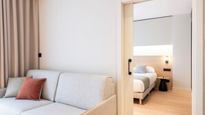 Una habitación de hotel moderna con un sofá de color claro, una cama con estructura de madera, una mesita redonda pequeña y un aplique de pared.