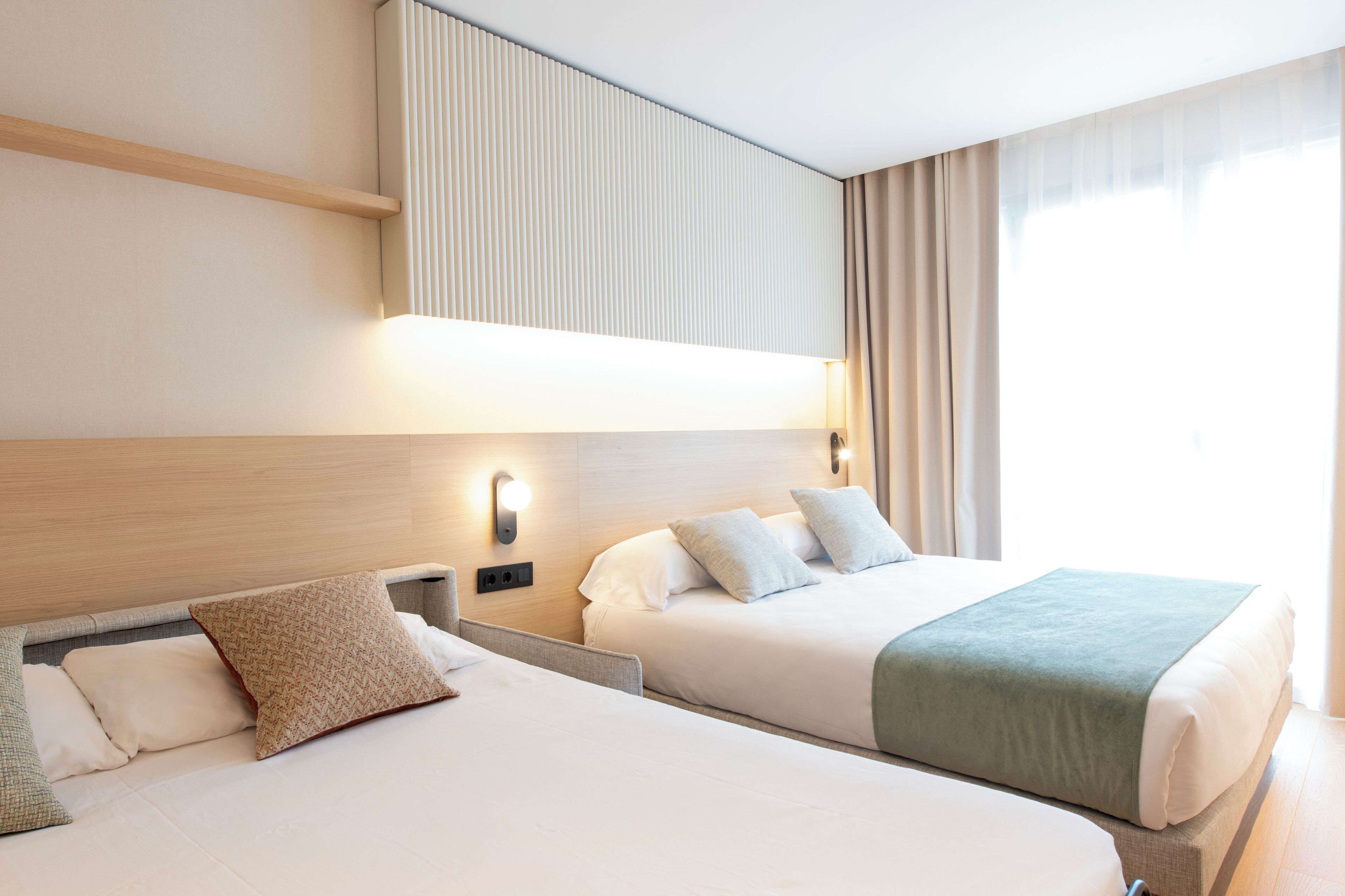 Habitación de hotel con dos camas, cabecera con iluminación integrada, ventana con cortinas y un aplique de pared.