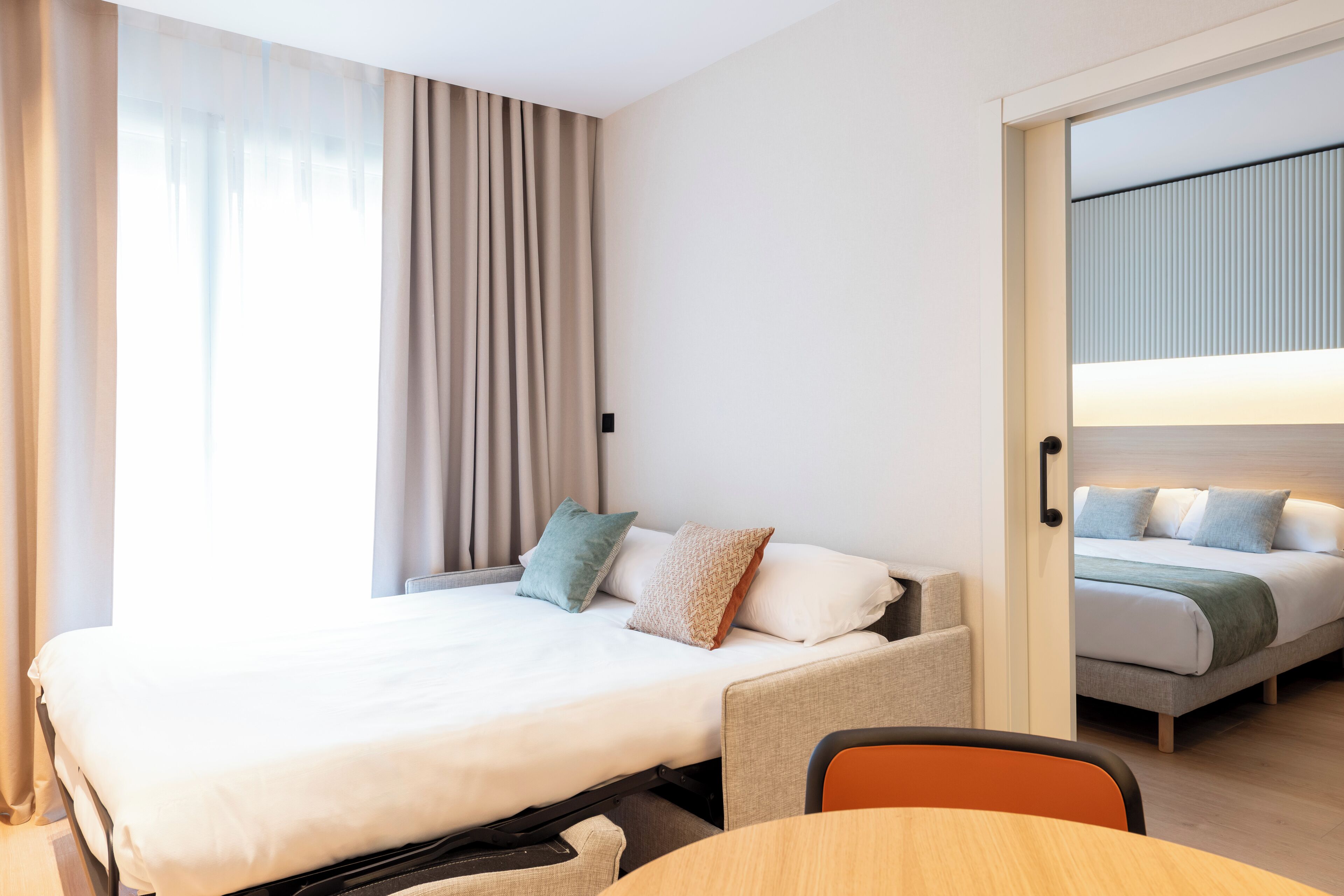 A hotel room with a bed, a chair, a table, and a view of another room.