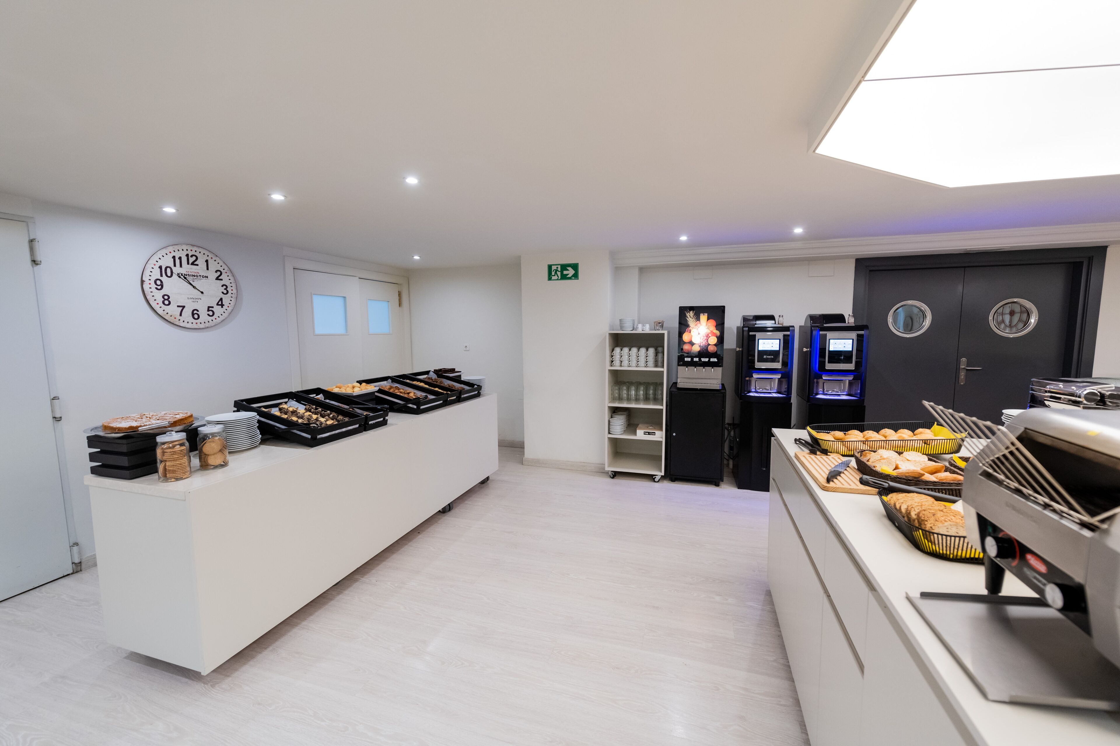 A modern hotel lobby with a reception desk, a clock, a coffee machine, and self-service beverage dispensers.