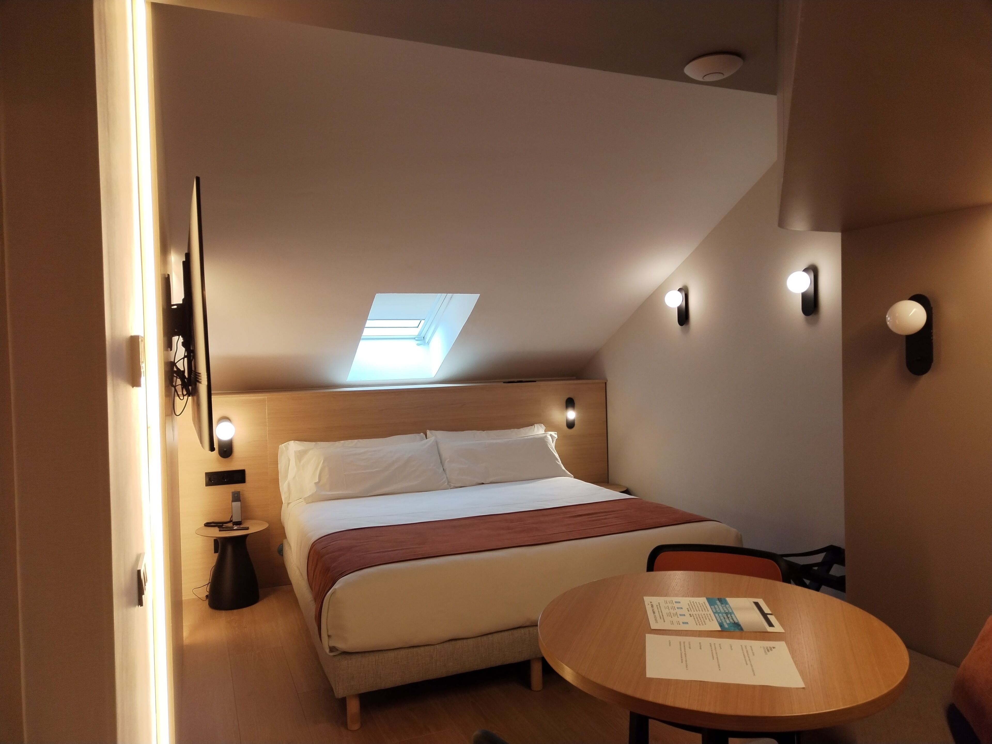 Ein Schlafzimmer mit einem Bett, einem runden Holztisch, einem kleinen Schreibtisch und einem Oberlicht.