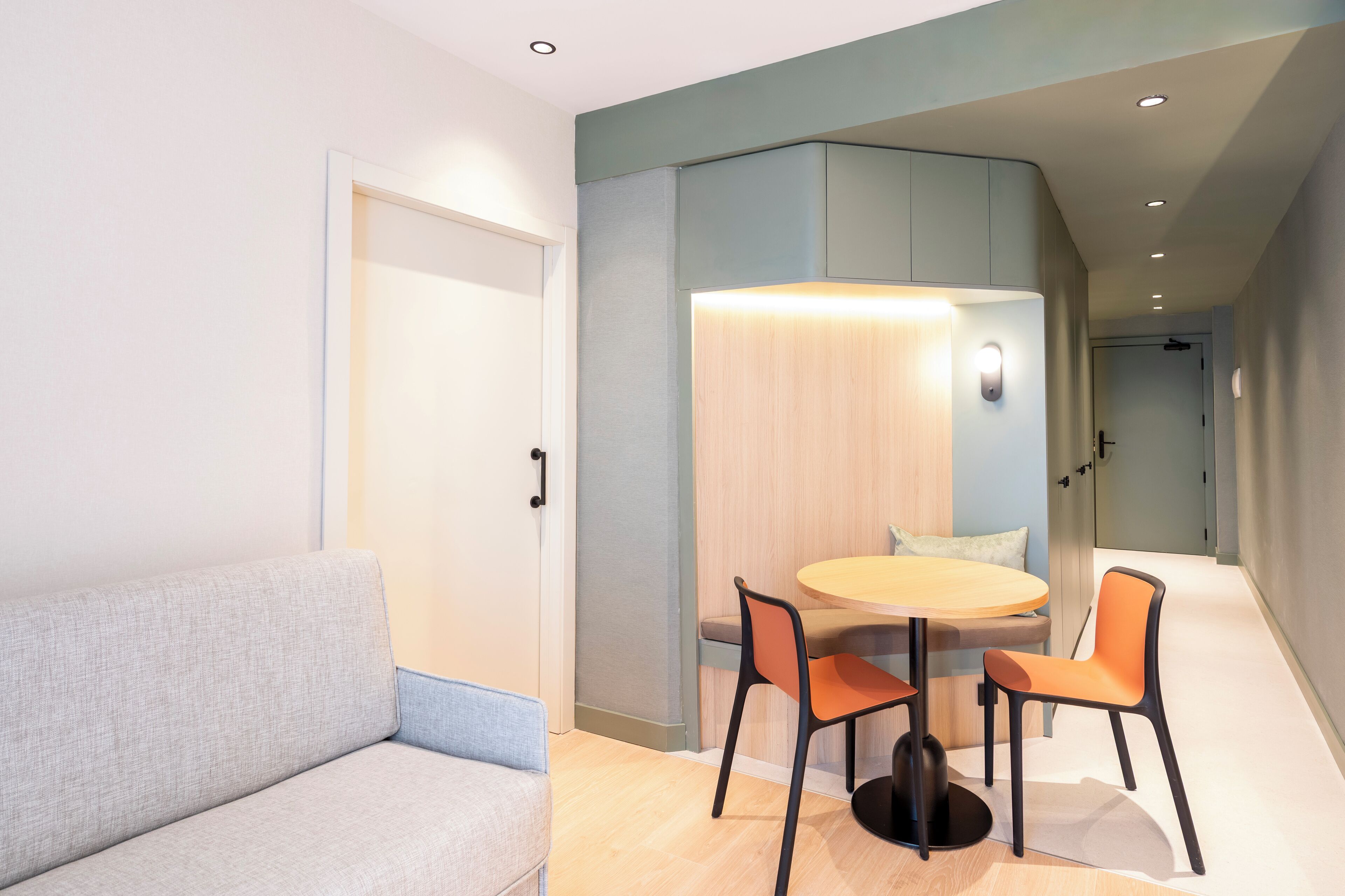 Ein modernes Hotelzimmer mit einer Couch, einem runden Tisch mit zwei Stühlen und einer hellen Wand mit einer eingelassenen Deckenleuchte.