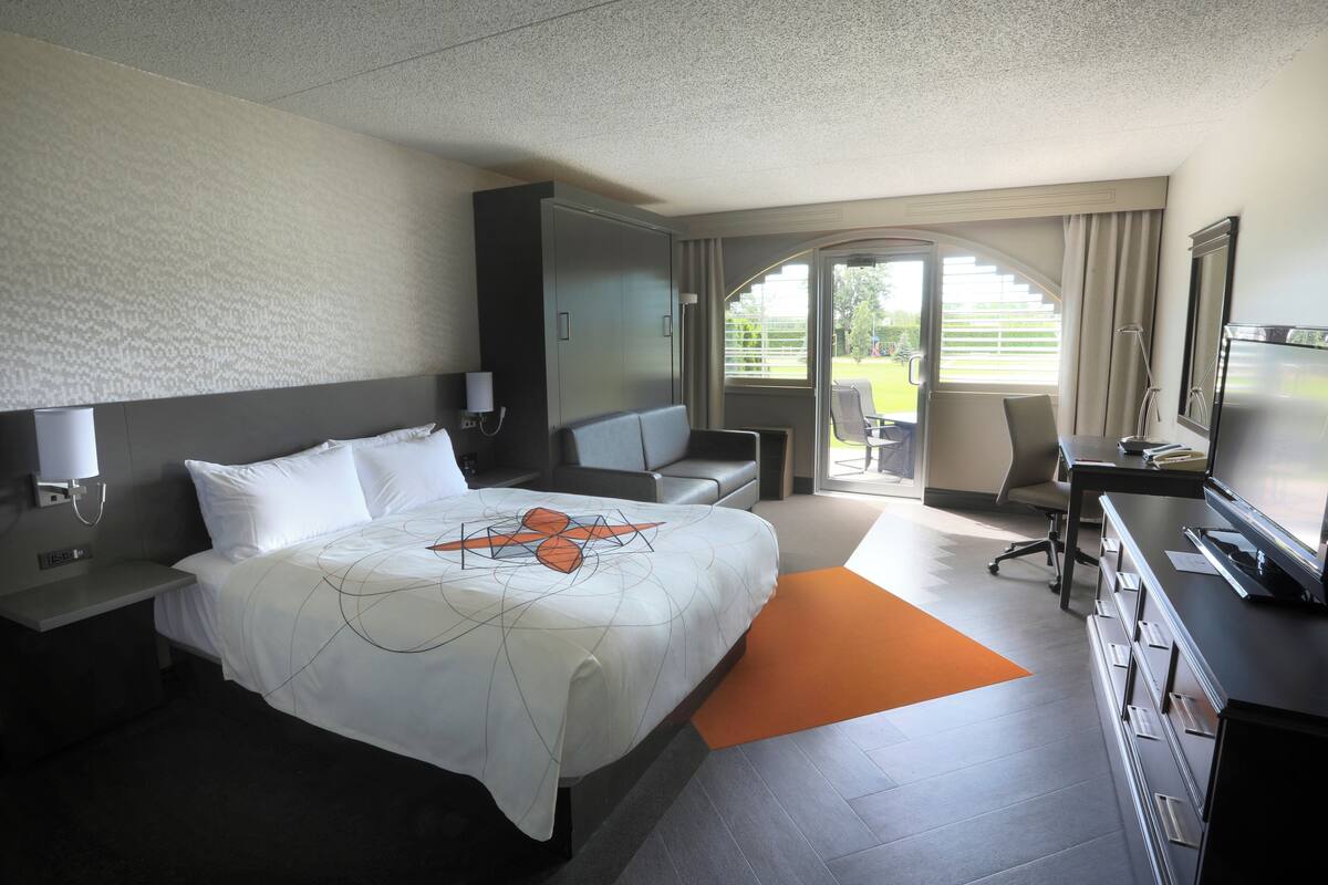 Quarto de hotel com uma cama grande, uma escrivaninha, uma cadeira, televisão e vista para o exterior.