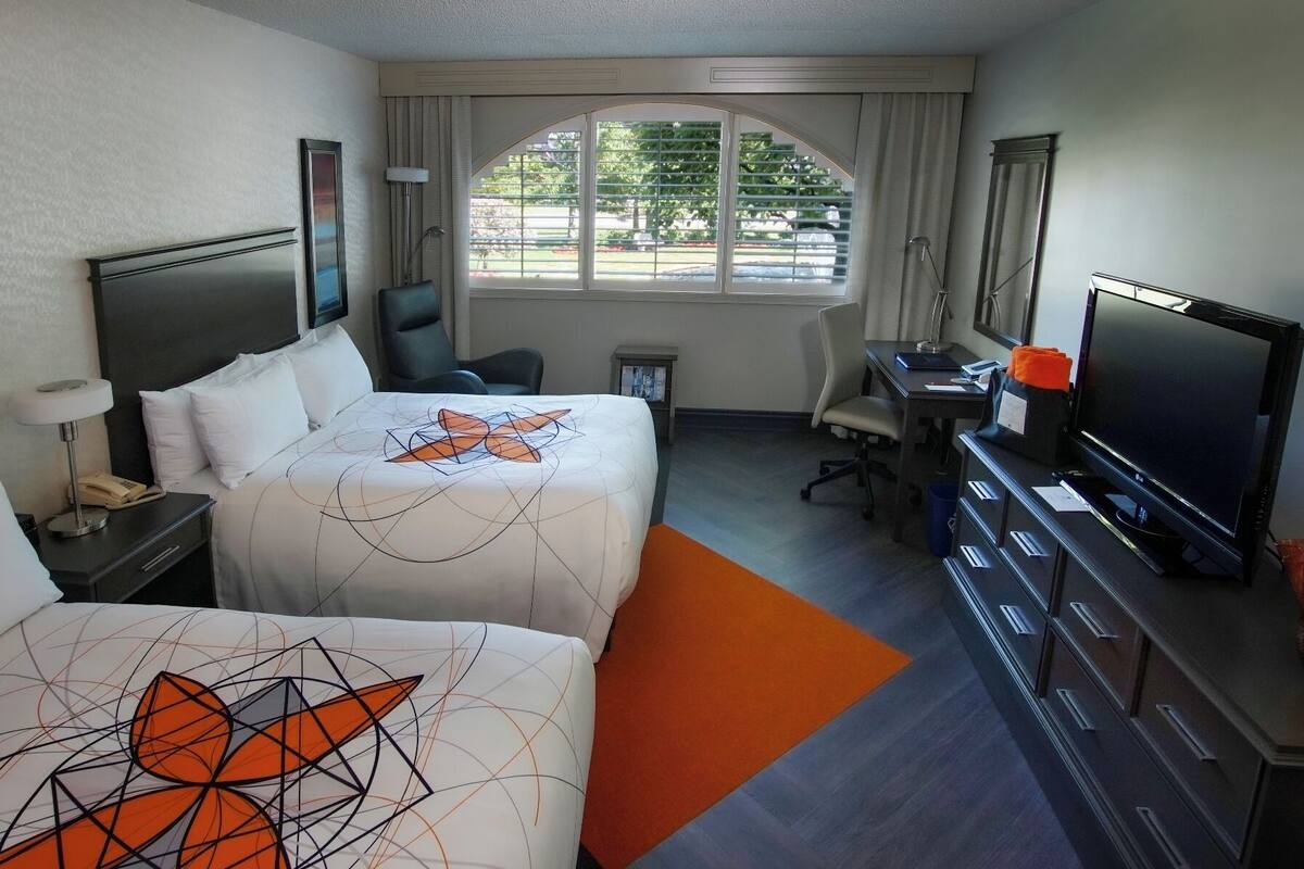Quarto de hotel com duas camas, uma mesa de trabalho com televisão, uma cadeira e uma janela com persianas.