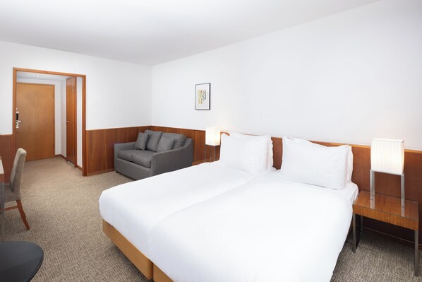 A hotel room with a large bed, a gray armchair, a wooden nightstand, and a desk with a chair.
