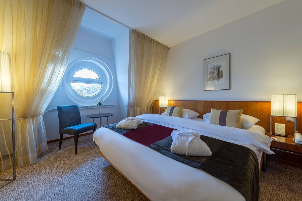 Une chambre d’hôtel avec un grand lit, une fenêtre ronde, une chaise et une table de chevet avec une lampe.