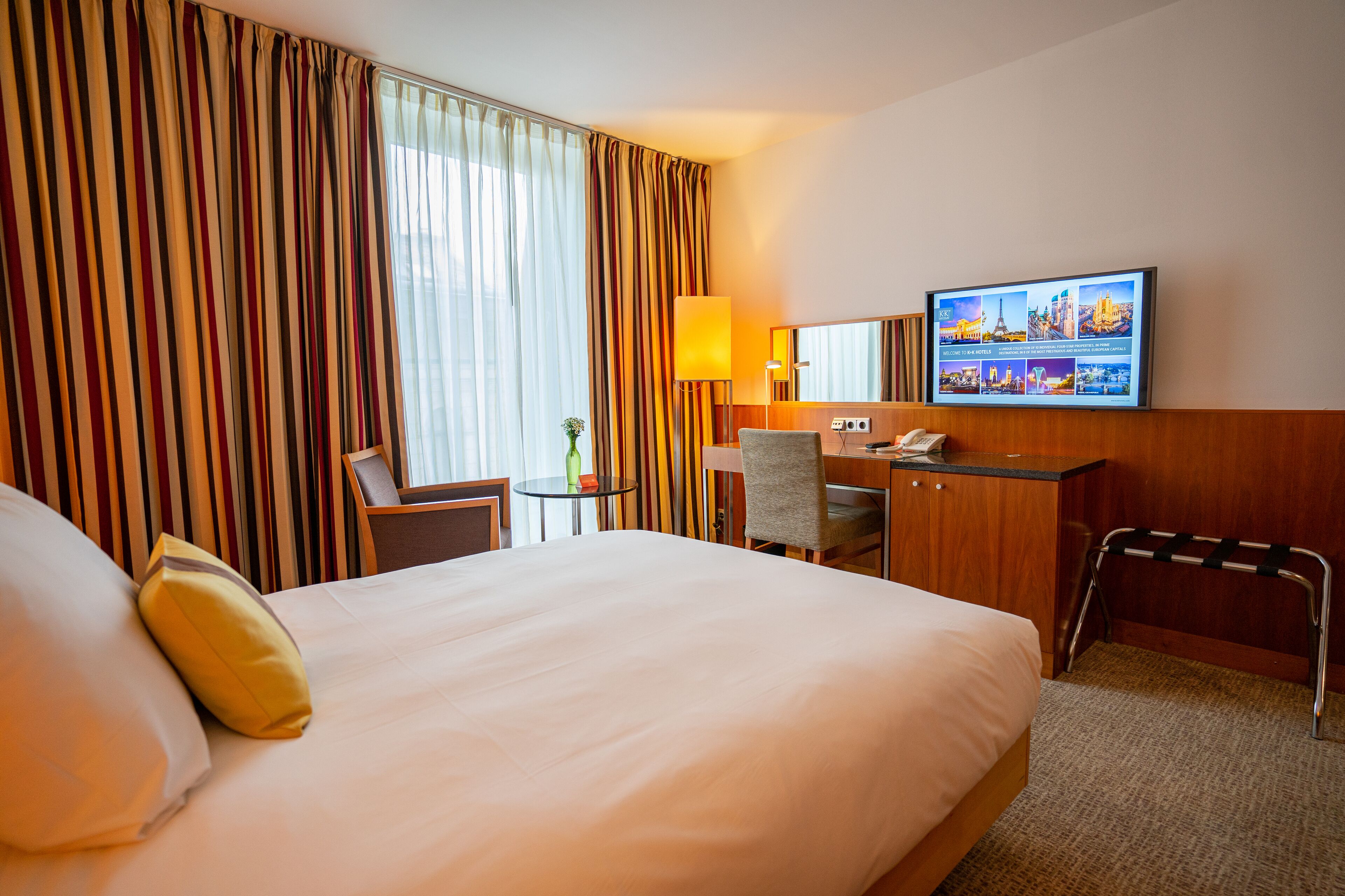 Ein Hotelzimmer mit einem großen Bett, einem Schreibtisch, einem Sessel, einem Fernseher und einem Fenster mit Vorhängen.