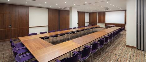 A conference room with a long wooden table, purple chairs, and a projector screen.