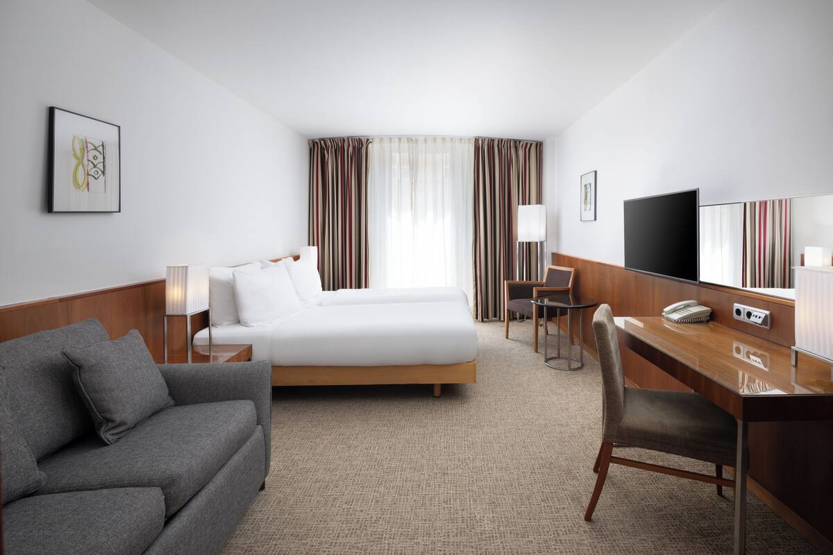 Une chambre d’hôtel avec un lit, un canapé gris, un bureau avec une chaise, une télévision et une fenêtre avec des rideaux.