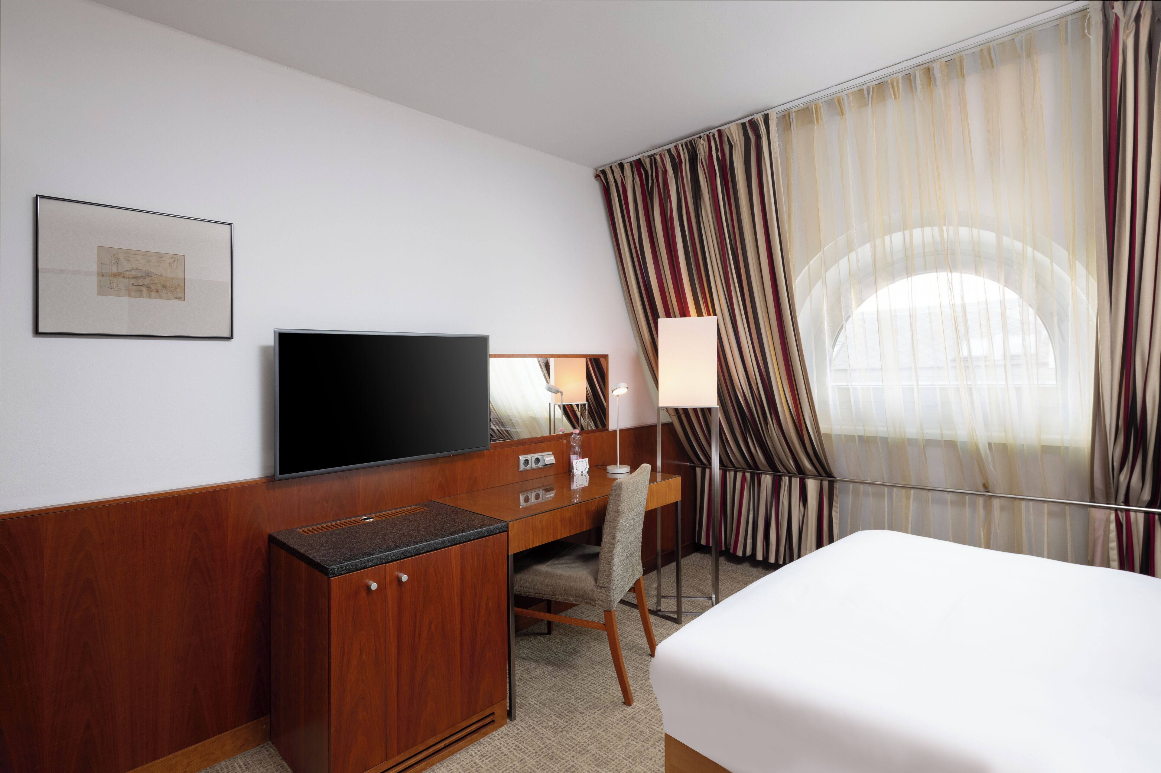 A hotel room with a bed, a desk with a flat-screen TV, a lamp, and a window with curtains.