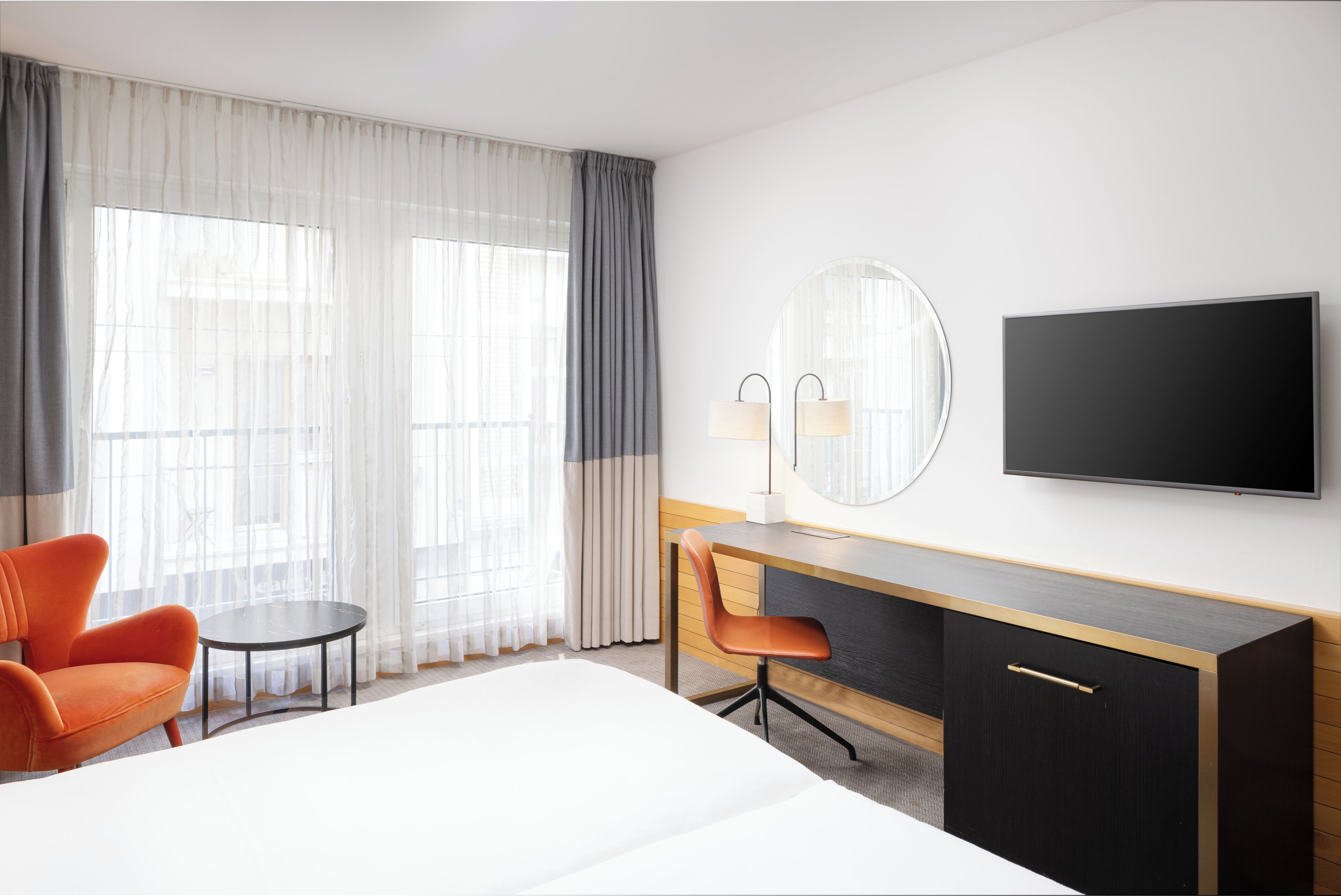 A hotel room with a bed, a desk, a chair, a TV, and large windows with curtains.