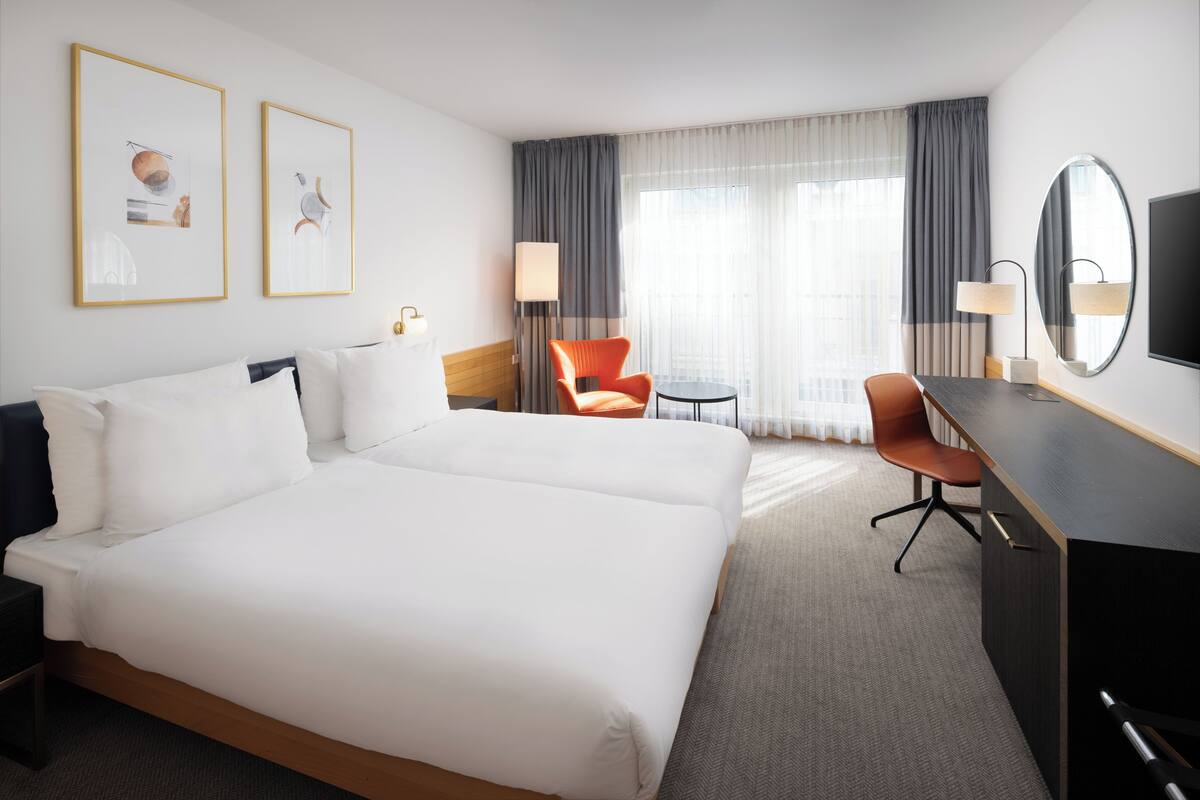 A hotel room with a large bed, two chairs, a desk, a TV, and a window with curtains.