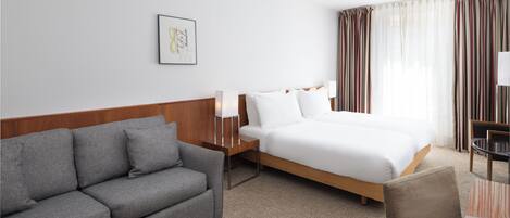 Una habitación de hotel con una cama, un sofá gris, una mesita y una lámpara.