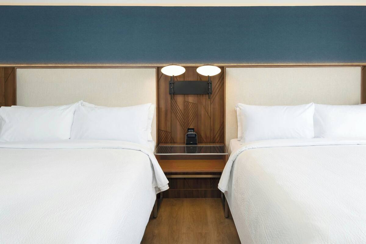 A hotel room with two beds, a wooden headboard, a nightstand with a clock, and a wall-mounted TV.