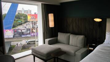 Habitación de hotel con sofá, reposapiés y cama. Se ve una ventana con vista a la calle y edificios.