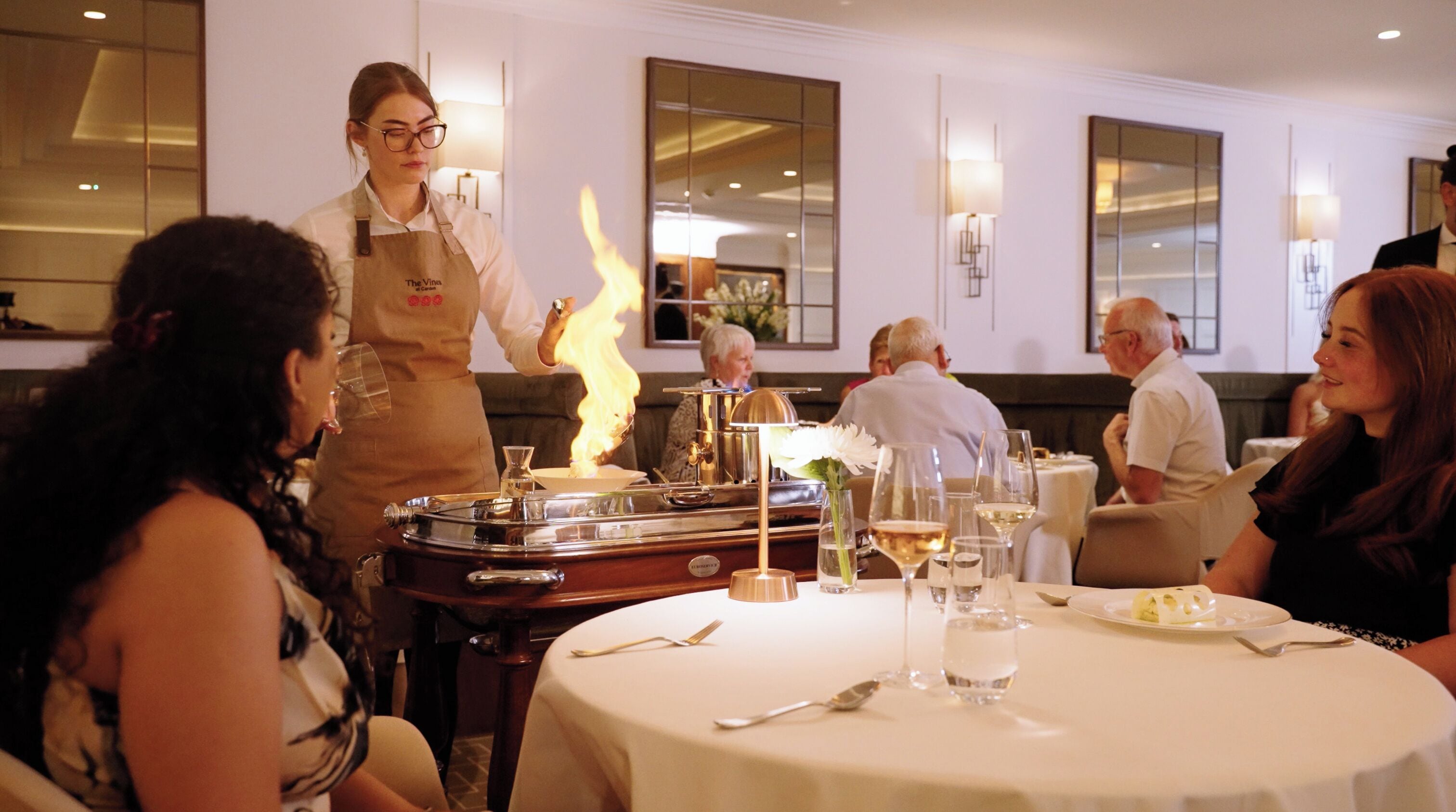 A dining area with round tables, a waitstaff, and a table with a lit stove.