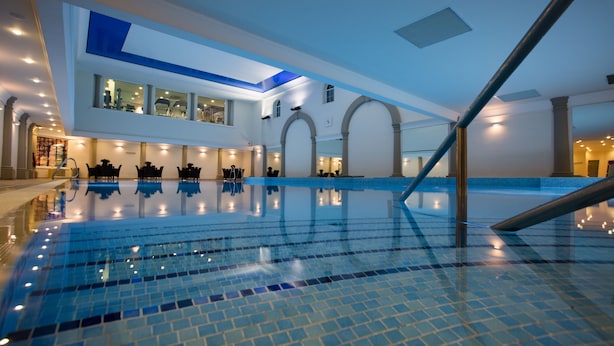 A large indoor swimming pool with clear blue tiles, a modern architectural design with arched columns, and a mezzanine level with seating. - Carden Park (Chester)