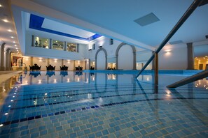 Una amplia piscina interior con azulejos azules cristalinos, diseño arquitectónico moderno con columnas en arco, y un entrepiso con asientos.