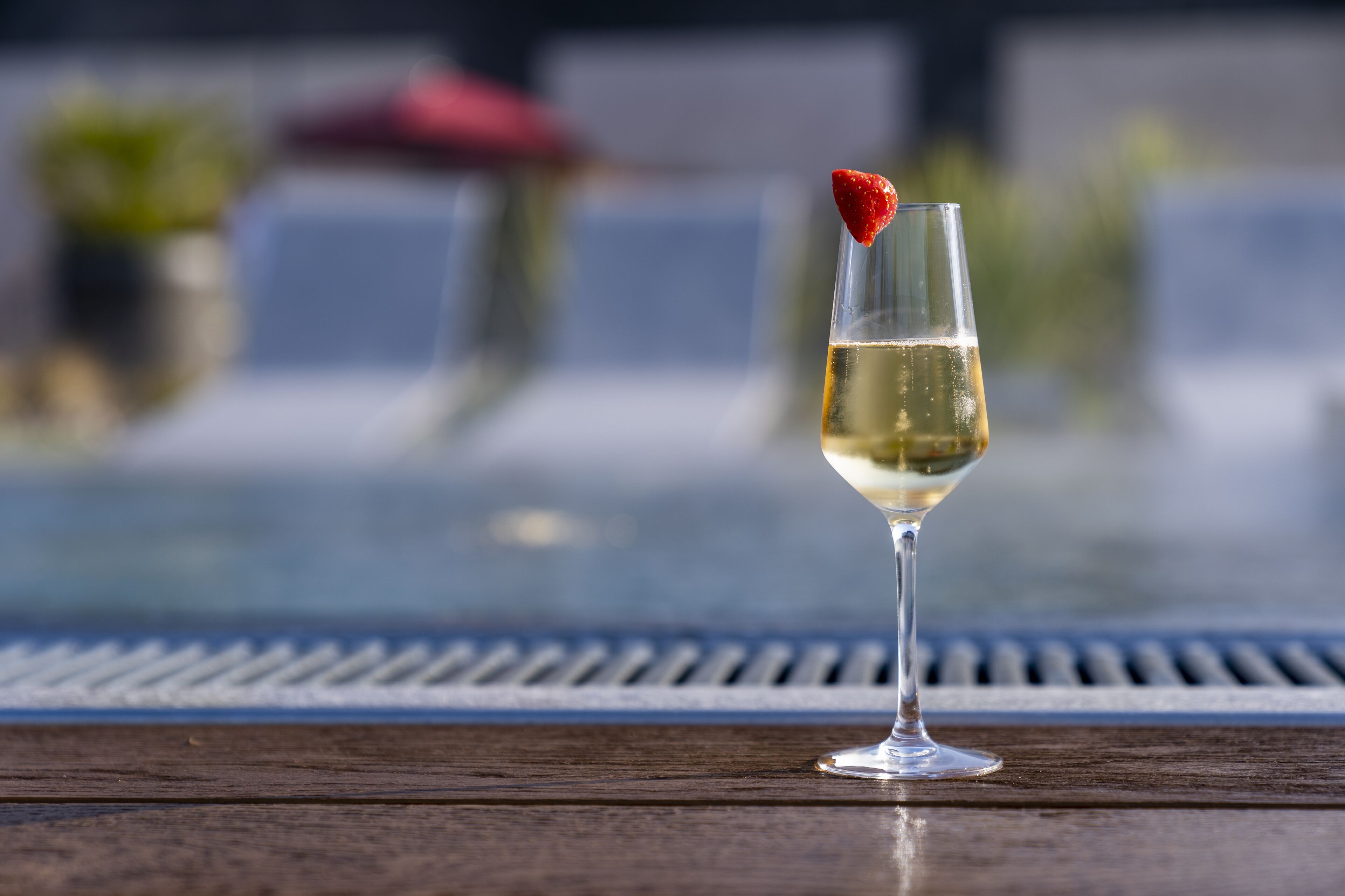 A glass of white wine with a strawberry garnish on a wooden table by a poolside.