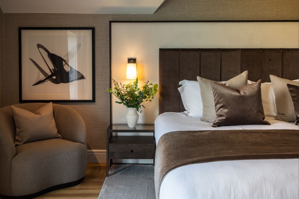 Ein modernes Hotelzimmer mit Bett, Nachttisch, Sessel, gerahmtem Kunstwerk und Wandlampe.