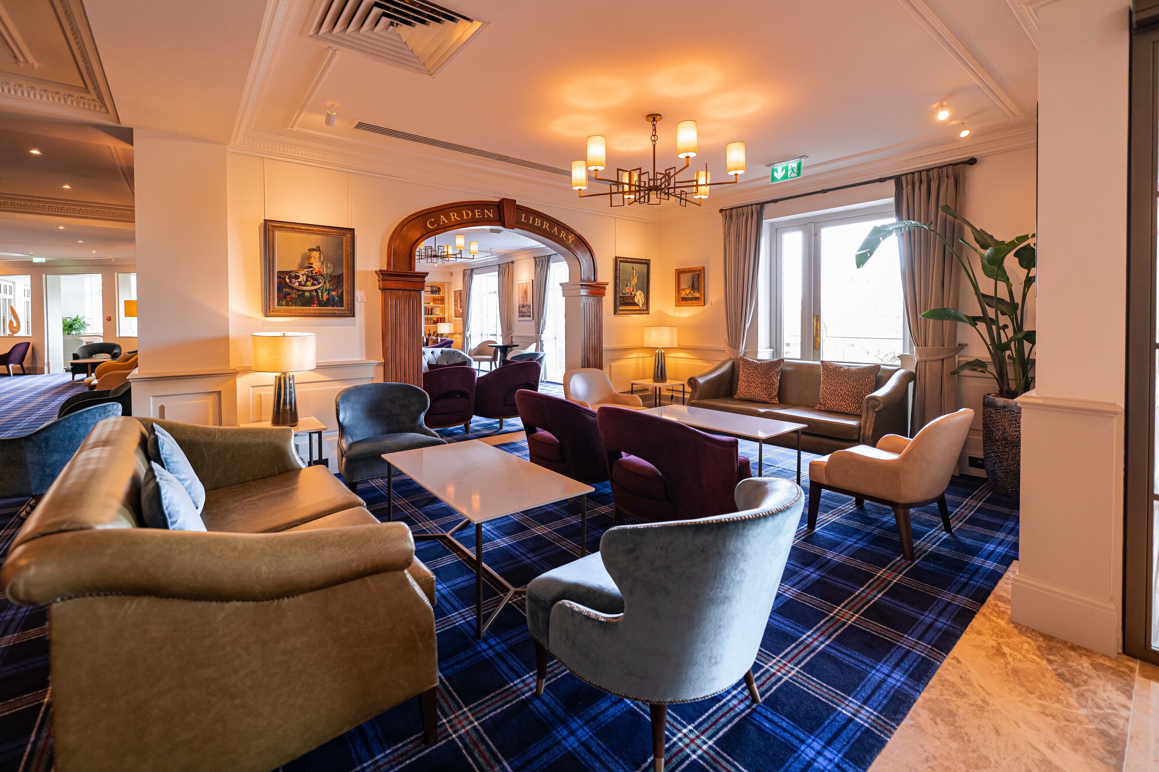 A lobby with a blue plaid carpet, leather sofas, and armchairs. There is a chandelier, a 'Carden Library' sign, and a large window with curtains.
