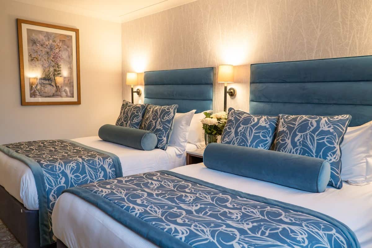 A hotel room with two beds, each with a blue headboard and matching pillows, a nightstand with a vase of flowers, and a framed picture on the wall.