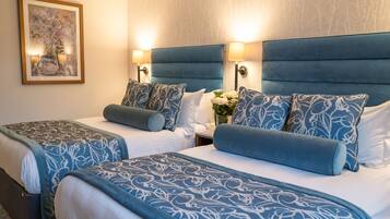 Une chambre d’hôtel avec deux lits, chacun doté d’une tête de lit bleue et d’oreillers assortis, une table de chevet avec un vase de fleurs et un tableau encadré au mur.