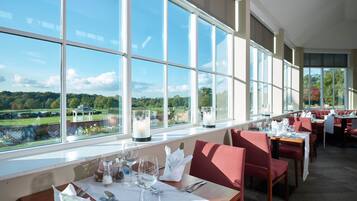 A restaurant with large windows offering a view of a green landscape. Tables are set with white tablecloths and red chairs.
