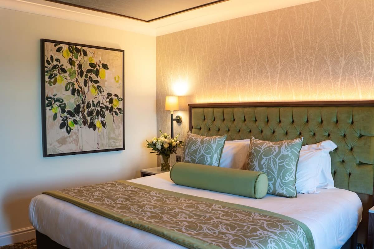 A hotel room with a large bed, a tufted headboard, a painting on the wall, and a nightstand with a lamp and flowers.