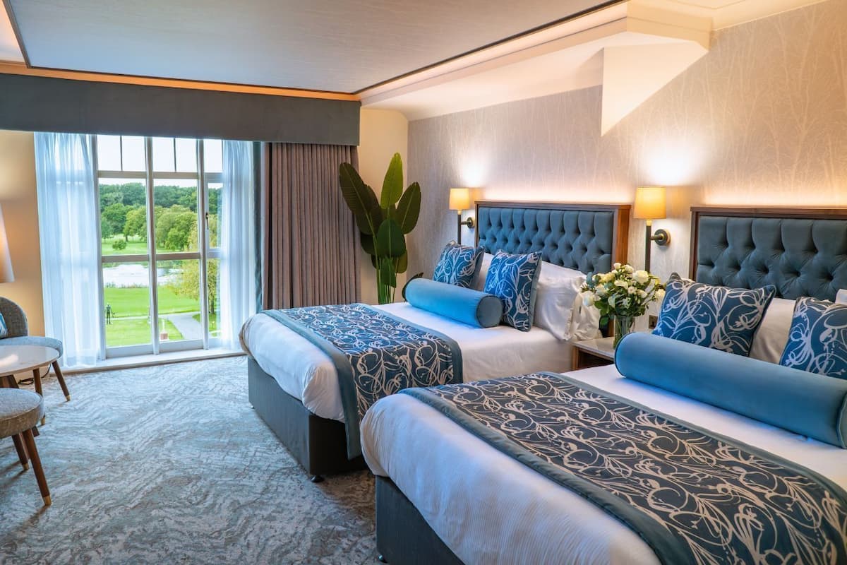 A hotel room with two beds, a large window with a view of a green landscape, a sitting area with a chair, and a plant.
