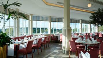 Un'ampia sala da pranzo con diversi tavoli e sedie, grandi finestre che offrono vista all'esterno e un design moderno del soffitto.