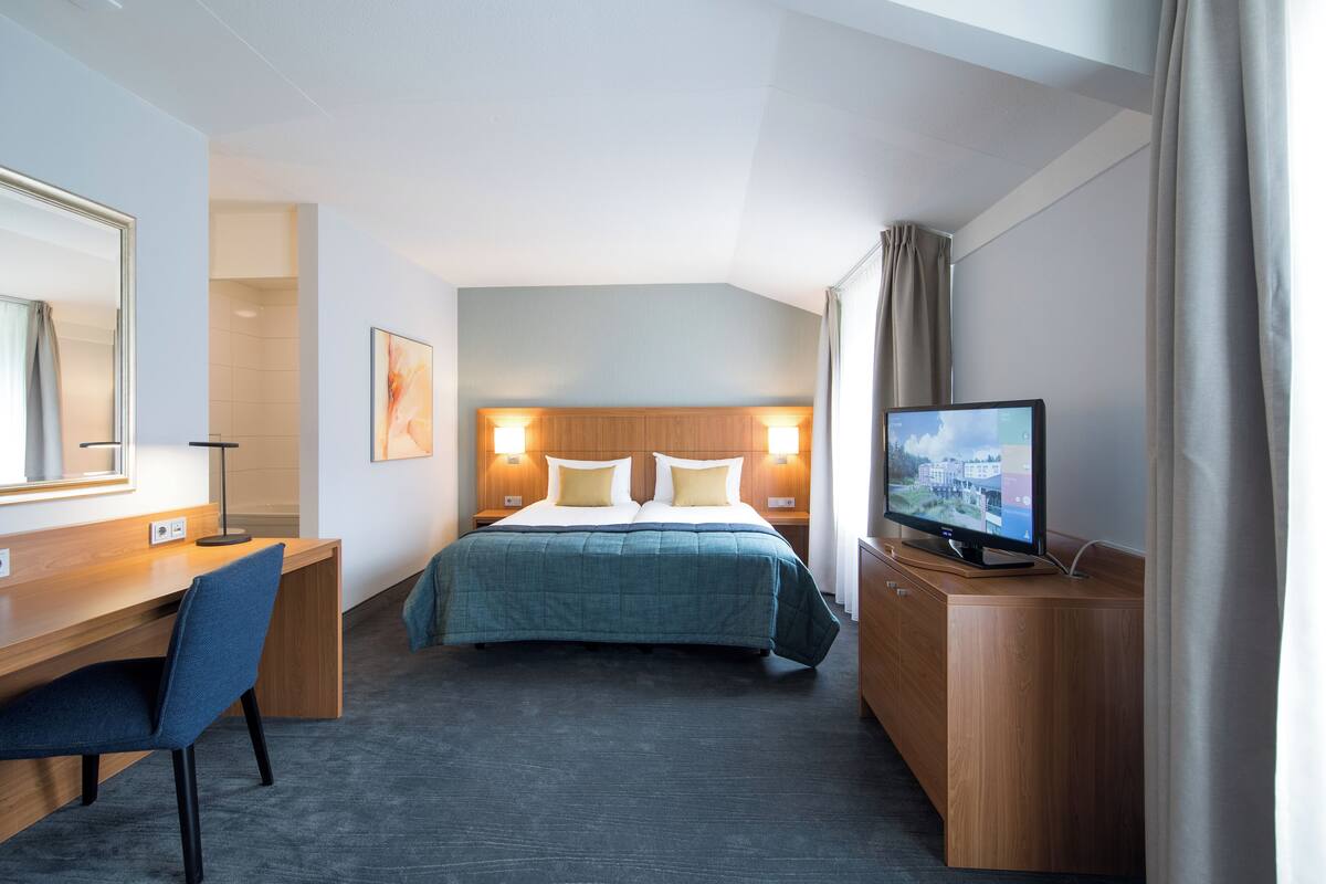 Habitación de hotel con cama, escritorio, silla, televisión y un amplio ventanal con cortinas.